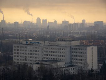 A photograph of a large concrete hospital with a blue SZPITAL sign against a hazy, smoggy city skyline with smoking chimneys