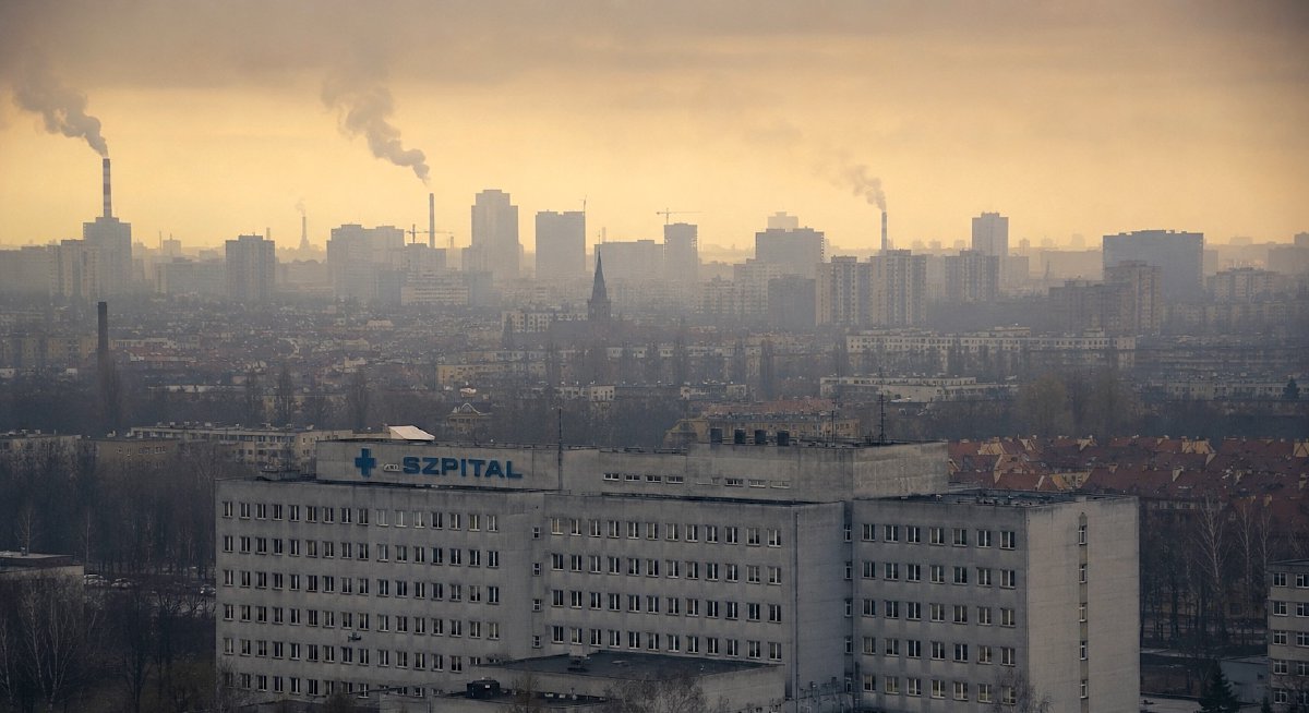 A photograph of a large concrete hospital with a blue SZPITAL sign against a hazy, smoggy city skyline with smoking chimneys